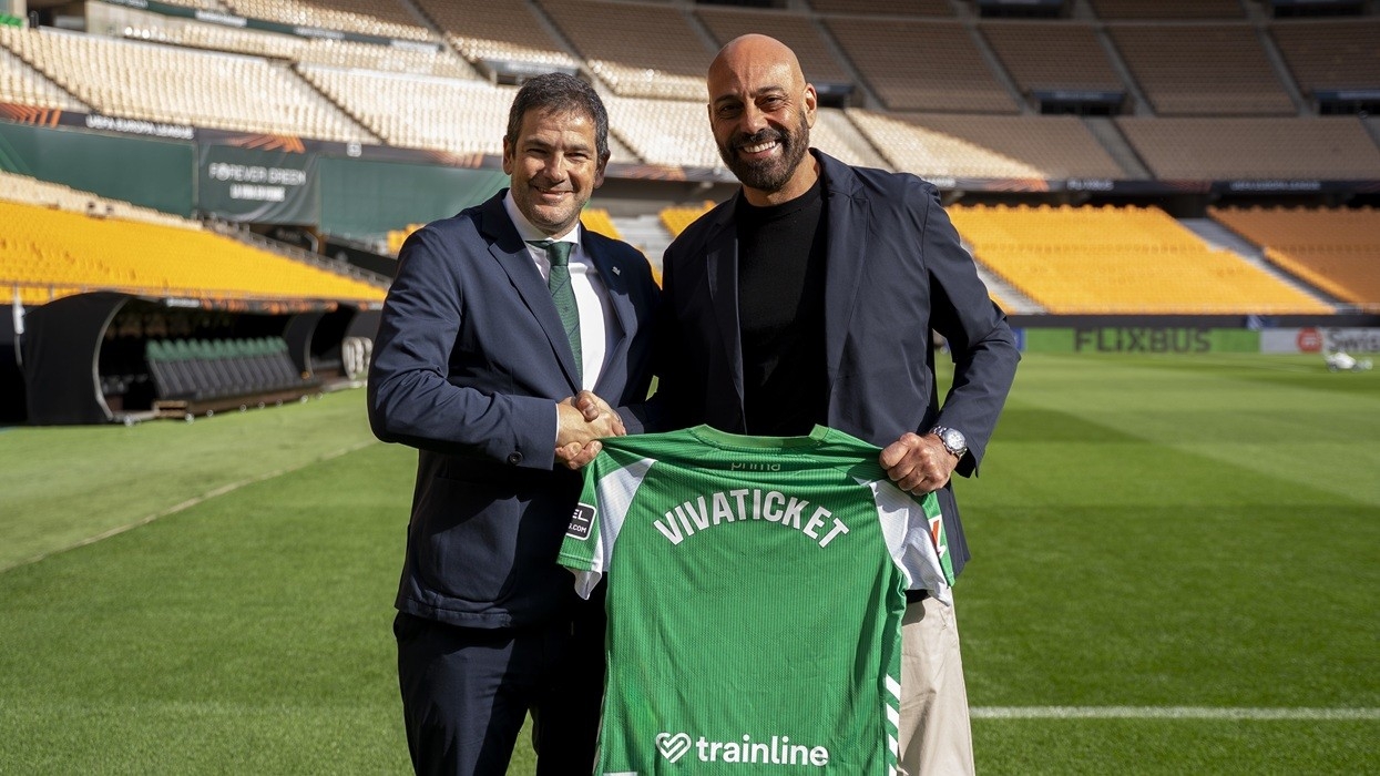 Two men smiling, holding a green jersey in an empty stadium.