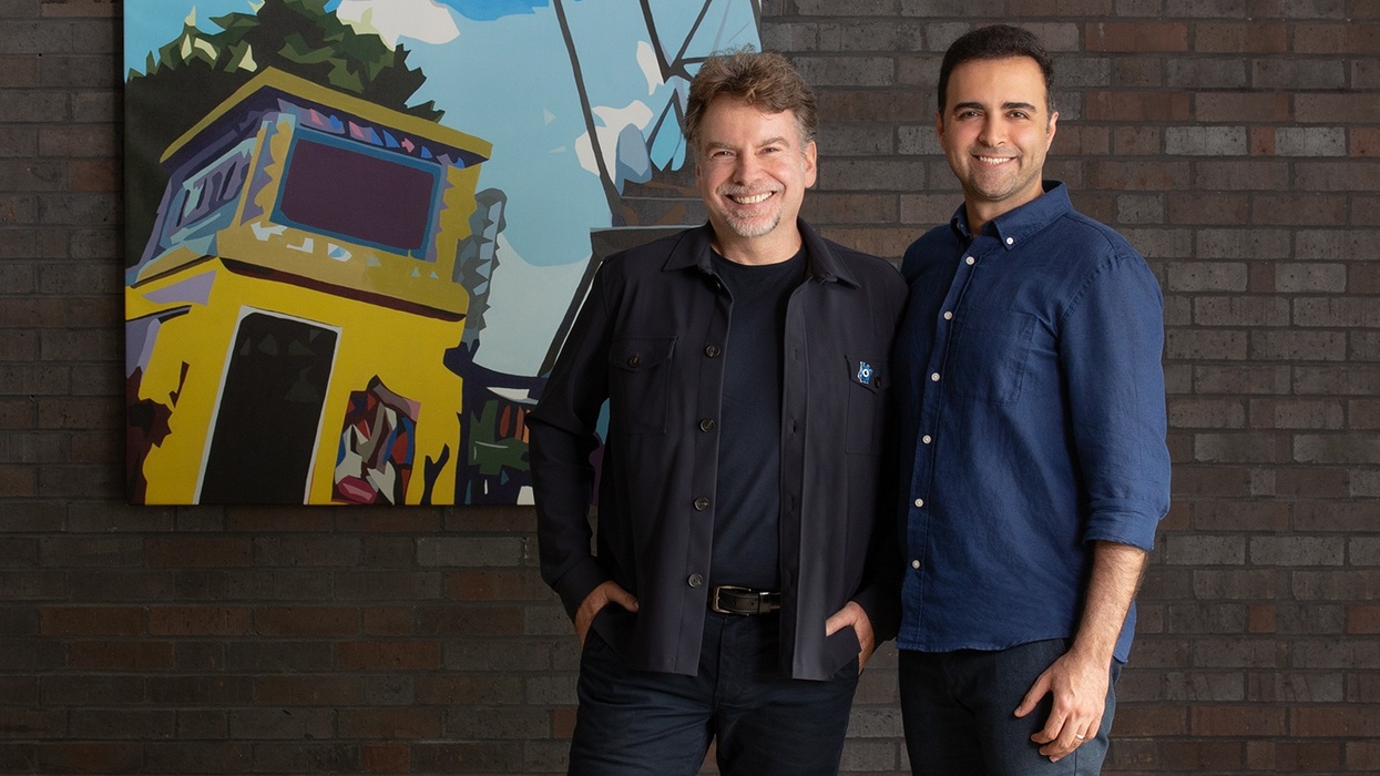 Two men smiling in front of a colorful abstract painting on a brick wall.