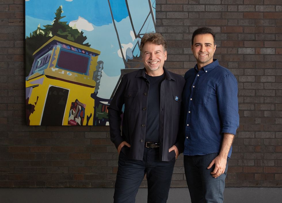 Two men smiling in front of a colorful abstract painting on a brick wall.
