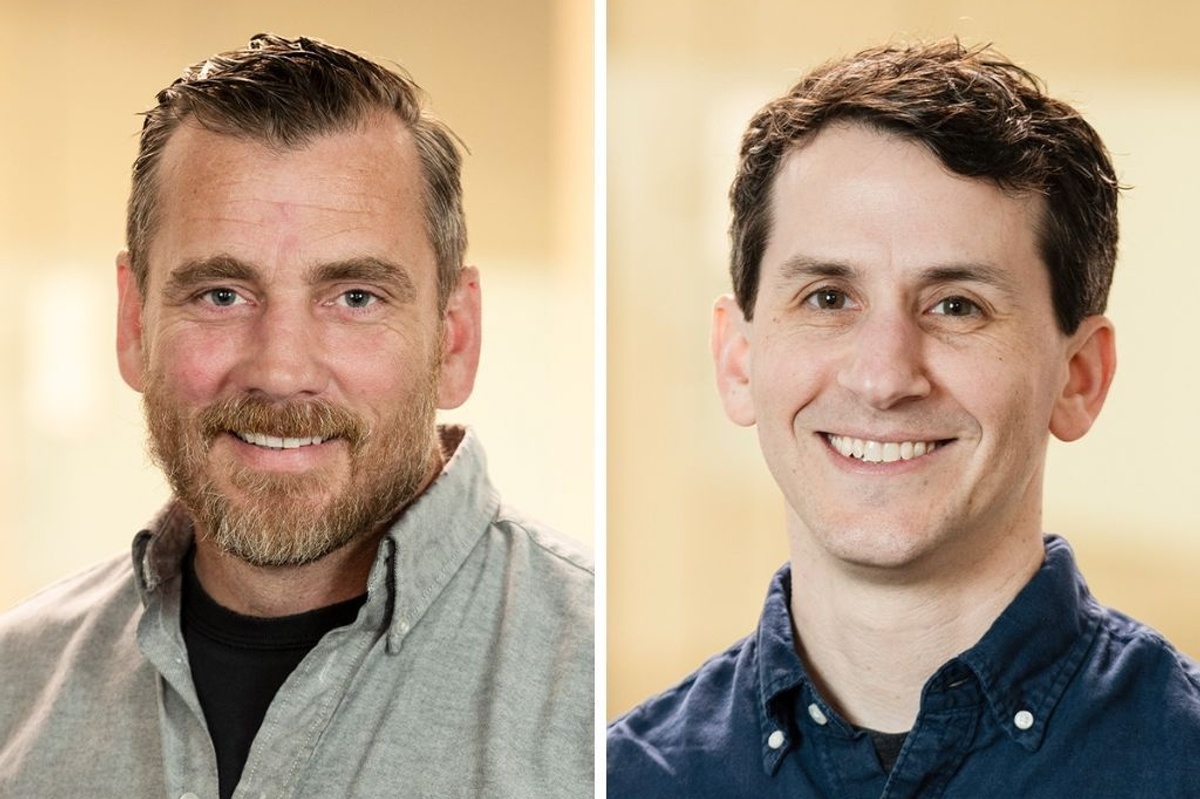Two men smiling, one with a beard and gray shirt, the other clean-shaven in a navy shirt.