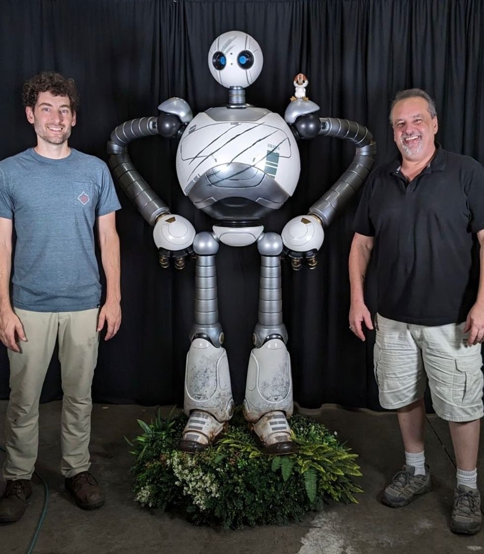 Two men stand beside a large, silver robot with a small figure on its shoulder.