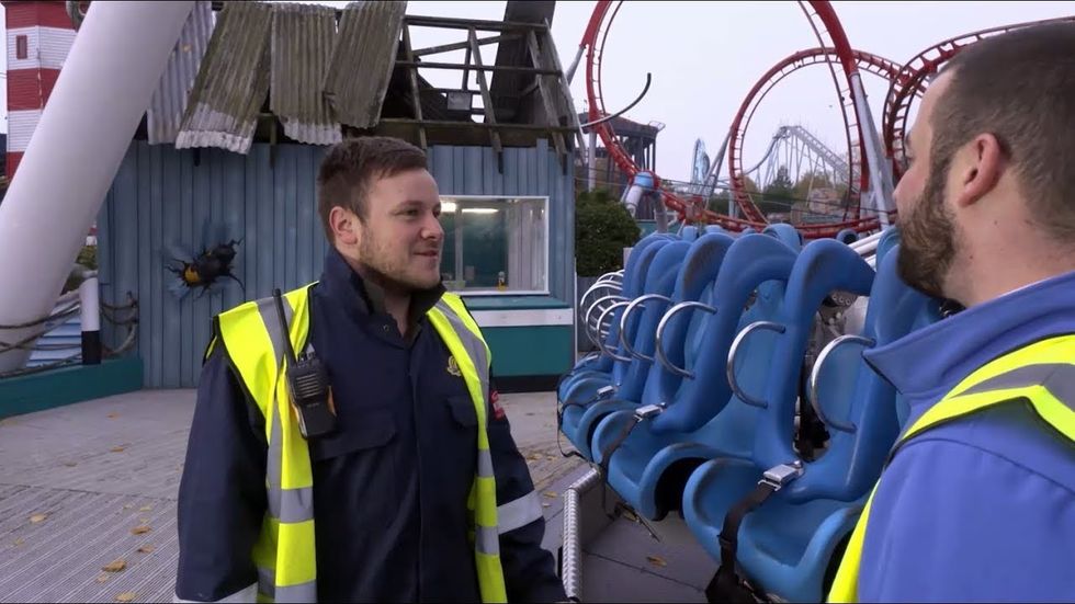Two men with high-vis vests talk near a roller coaster at an amusement park.