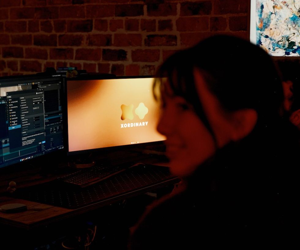 Two monitors on a desk with a blurred person in the foreground, brick wall backdrop.