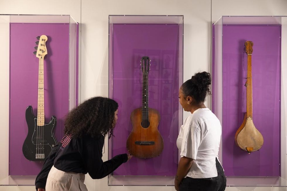 Two people admire guitars and a tanpura in a museum display with purple backgrounds.