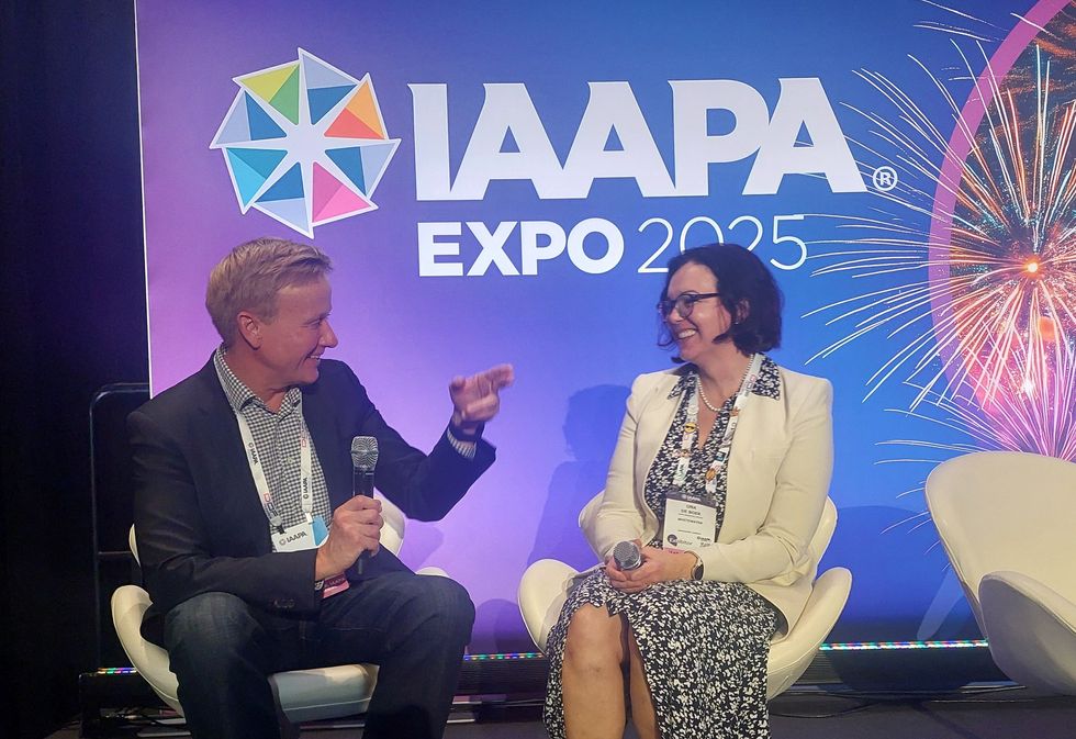 Two people chat on stage at IAAPA Expo 2025, with fireworks in the background.