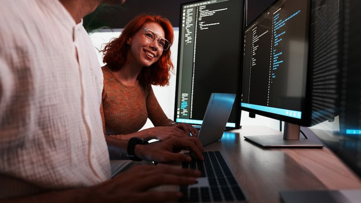 Two people coding on laptops, with multiple screens displaying code in the background.