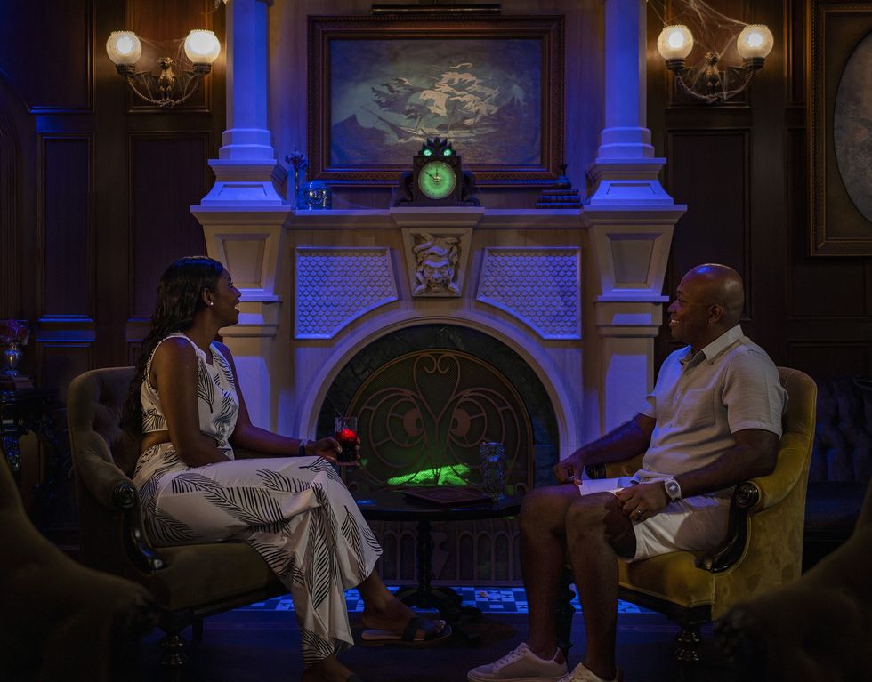 Two people conversing by a lit fireplace in a dim, elegantly decorated room.