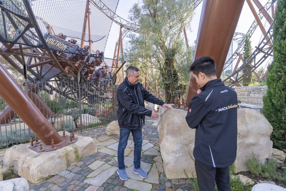 Two people discussing near a roller coaster with riders in the background.