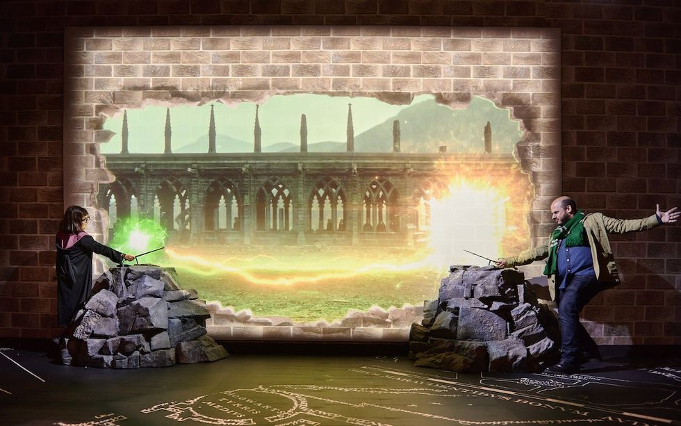 Two people duel with magic wands in front of a large screen with castle ruins.