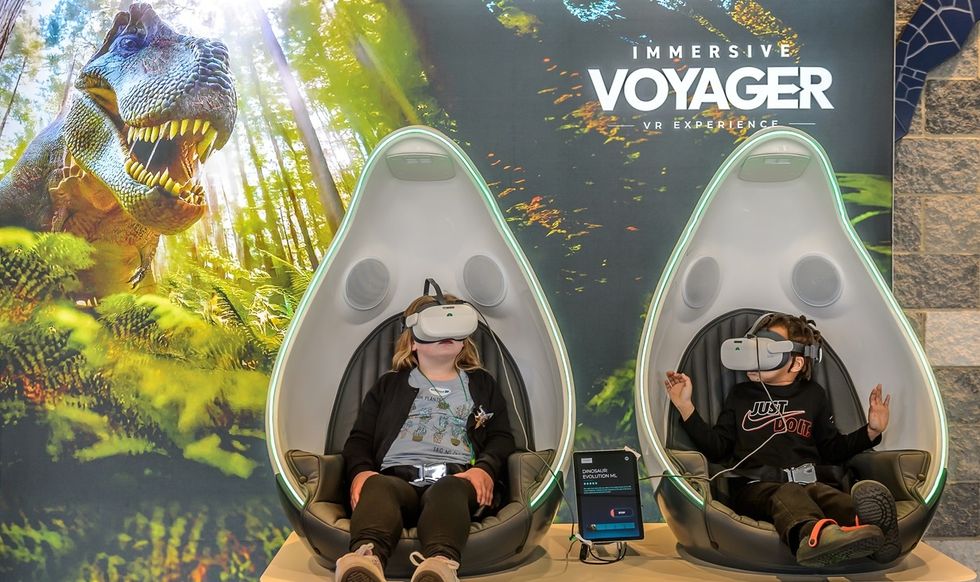 Two people enjoy a VR dinosaur experience in futuristic chairs.
