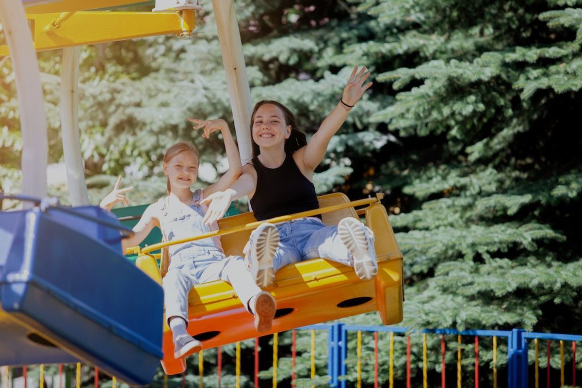 Two people enjoying a swing ride, smiling and waving, with trees in the background.