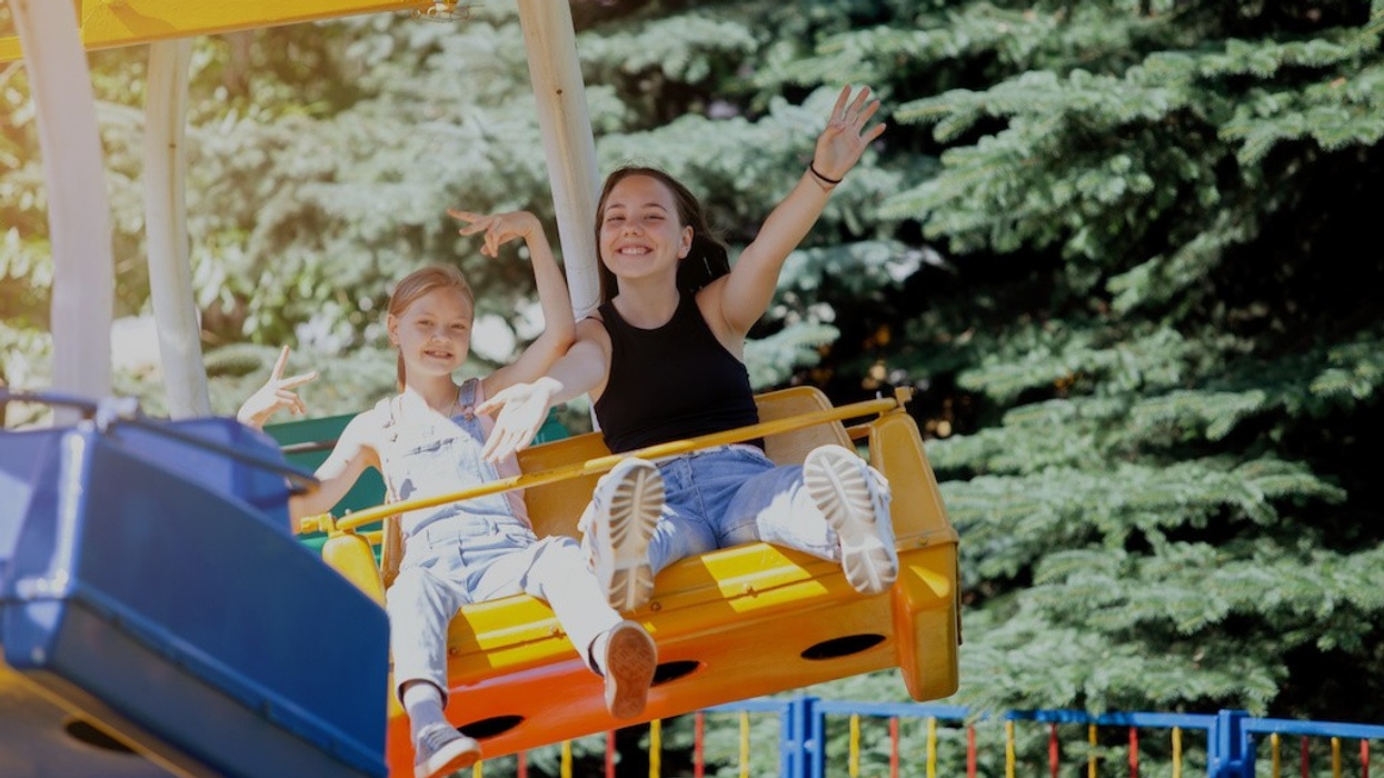 Two people enjoying a swing ride, smiling and waving, with trees in the background.