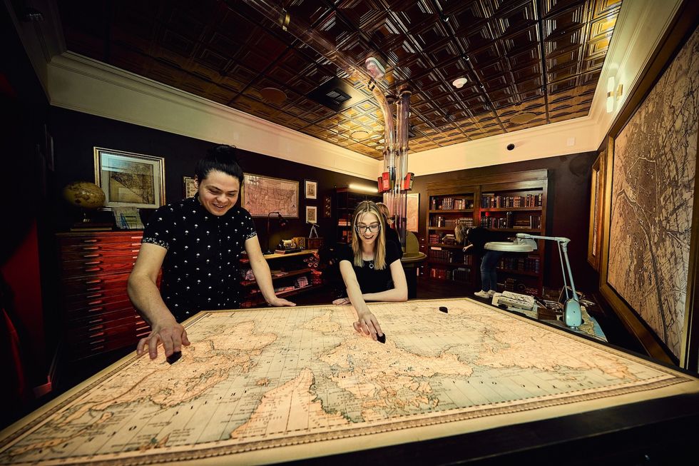 Two people explore a large vintage map in a cozy, book-filled room.