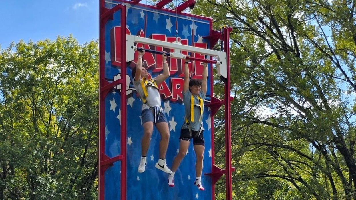 Two people holding onto pull-up bars on a vertical wall challenge outdoors.