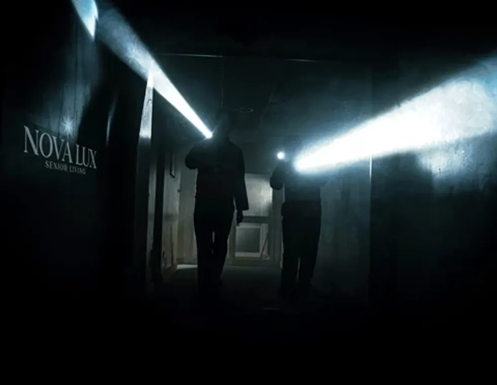 Two people in a dark hallway with flashlights, near a "Nova Lux Senior Living" sign.