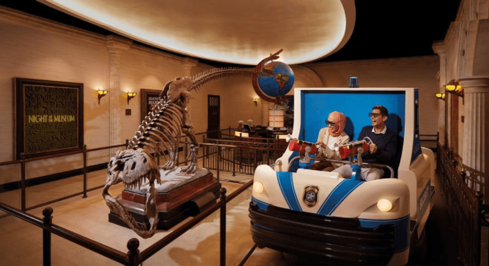 Two people in a museum ride car, passing a dinosaur skeleton exhibit.