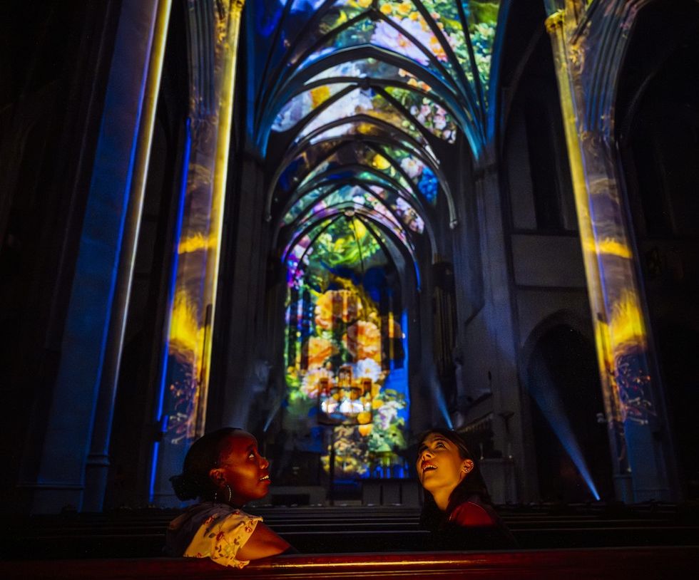 Two people in Grace Cathedral for AURA, colorful lights projected on the vaulted ceiling.
