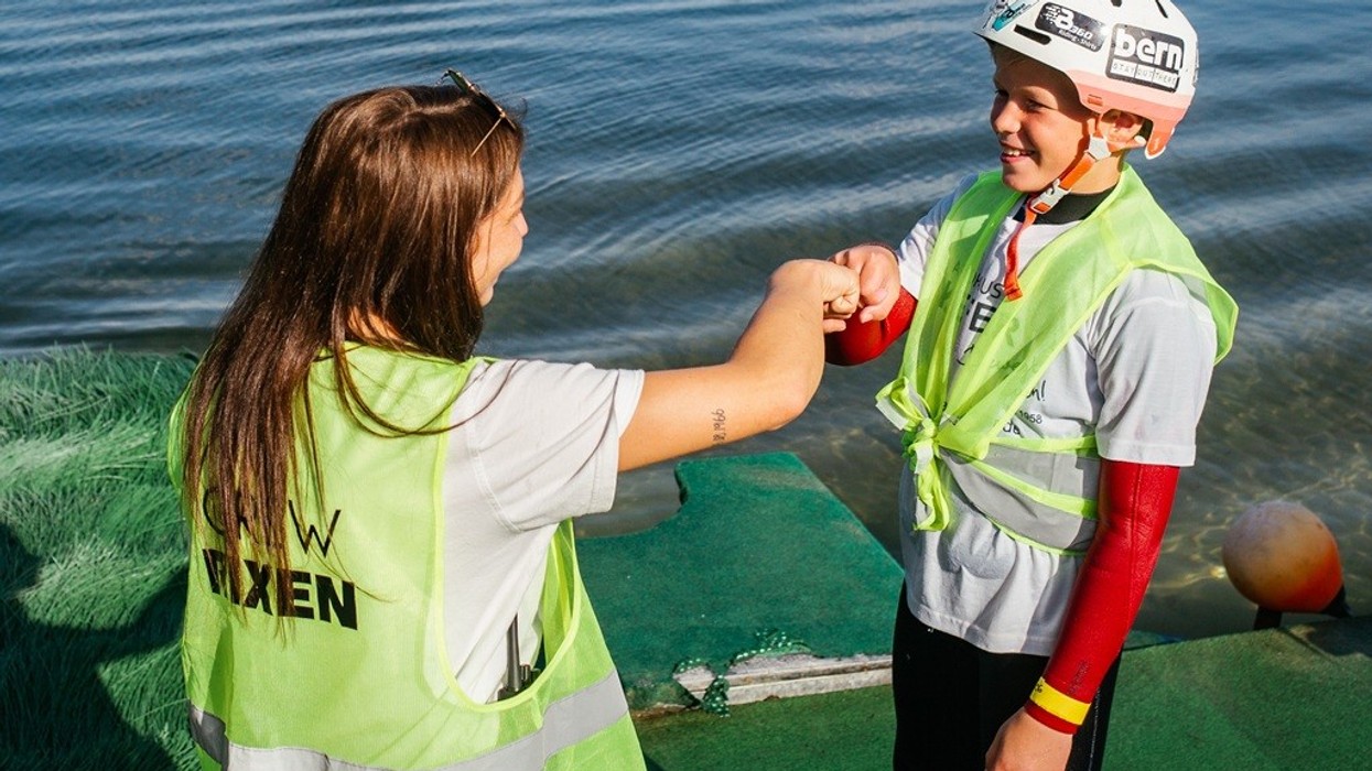 Two people in life vests fist-bump on a lakeside platform.
