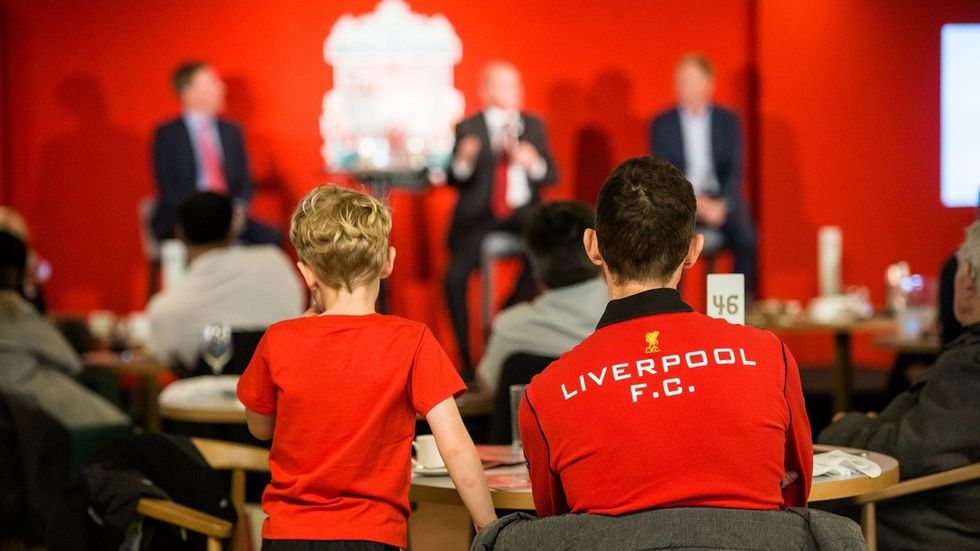 Two people in Liverpool F.C. shirts watch a panel discussion in a red-themed room.