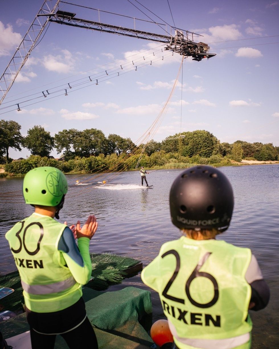Two people in neon vests watch a wakeboarder on a cable over a lake.