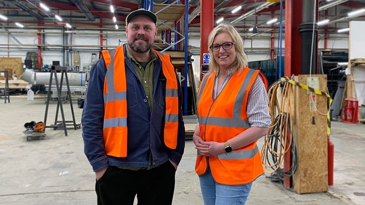 Two people in safety vests stand in an industrial warehouse setting, smiling.