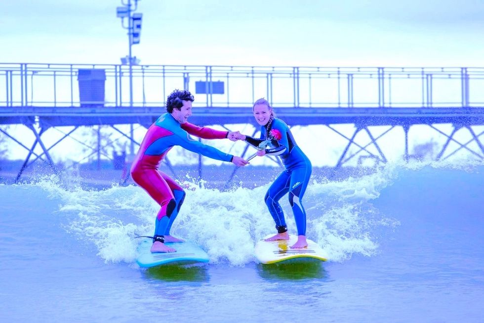 Two people in wetsuits surfing together, holding hands, with a pier in the background.