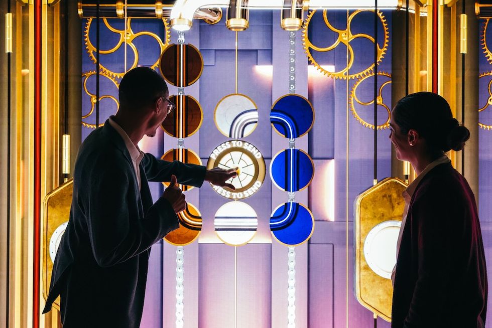 Two people interact with a colorful, mechanical display with gears and circular elements.