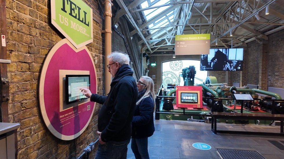 Two people interact with a touchscreen in an industrial-themed exhibit area.