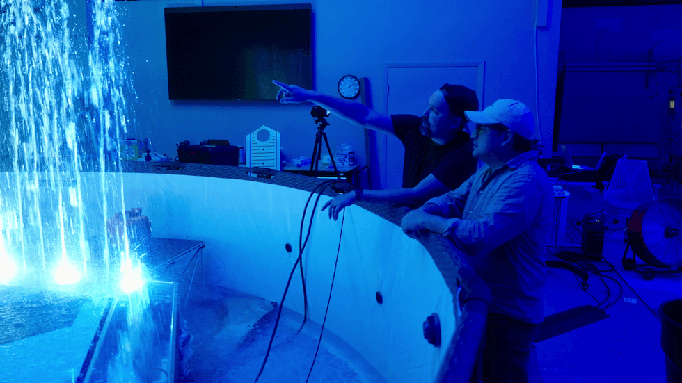 Two people observe and point at a lit water fountain in a blue-lit room.