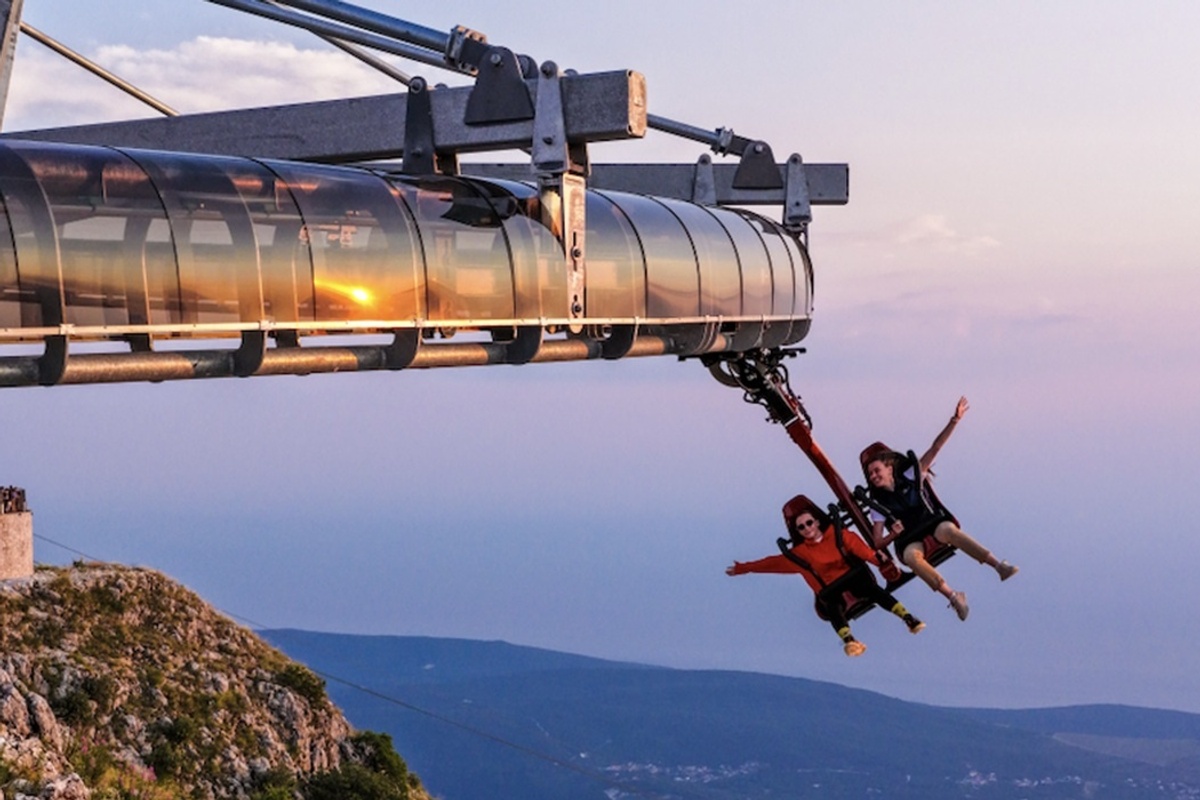 Two people on a Wie-Flyer at Lovćen National Park over a scenic landscape at sunset, joyfully spreading their arms.