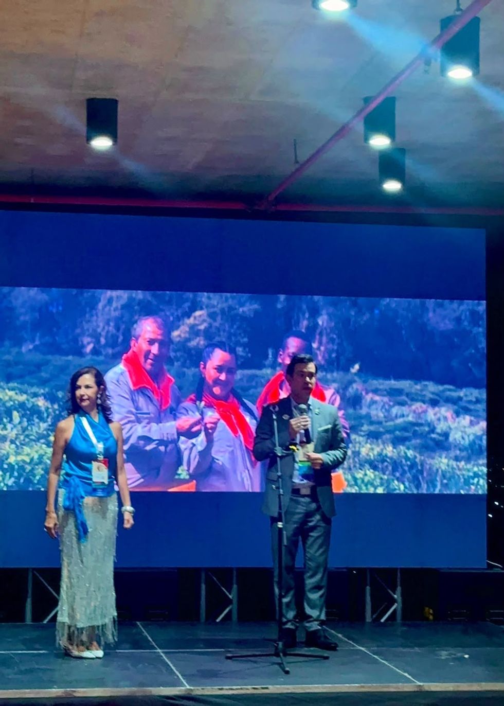 Two people on stage with a large screen backdrop showing three individuals outdoors.