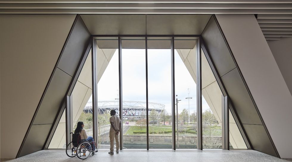 Two people, one in a wheelchair, gaze through large windows inside V&A East Museum overlooking a stadium.