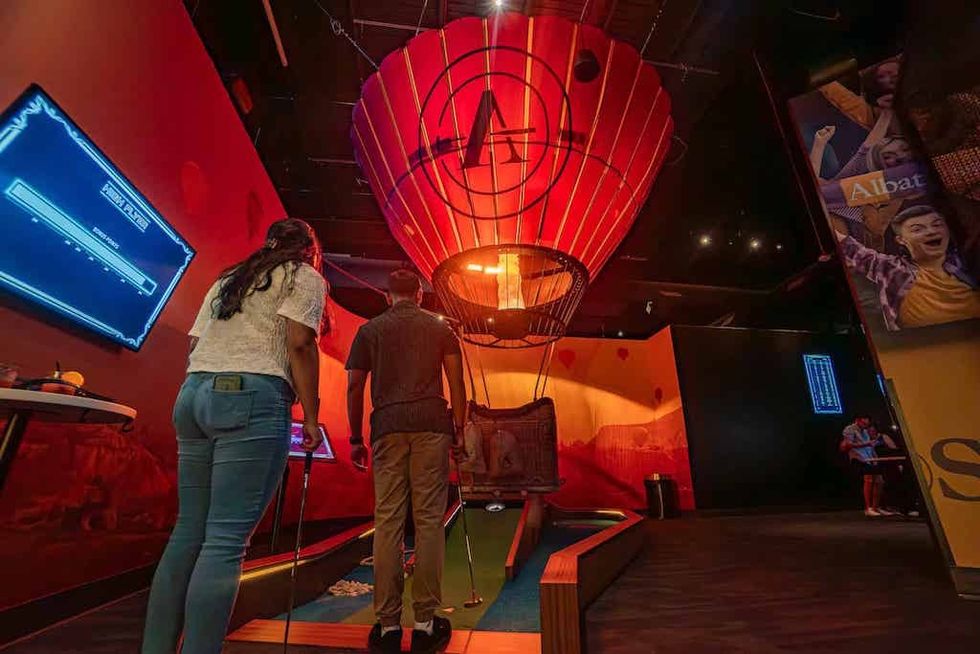 Two people play mini-golf indoors under a large, illuminated hot air balloon structure.