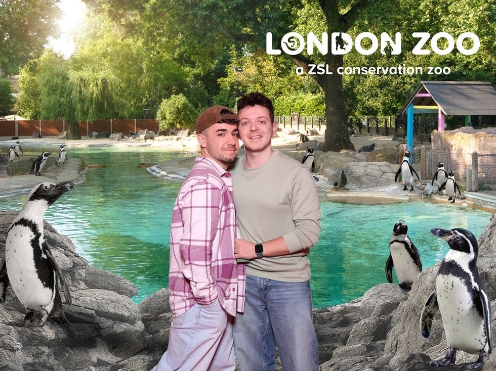 Two people pose at London Zoo, surrounded by penguins near a pool.