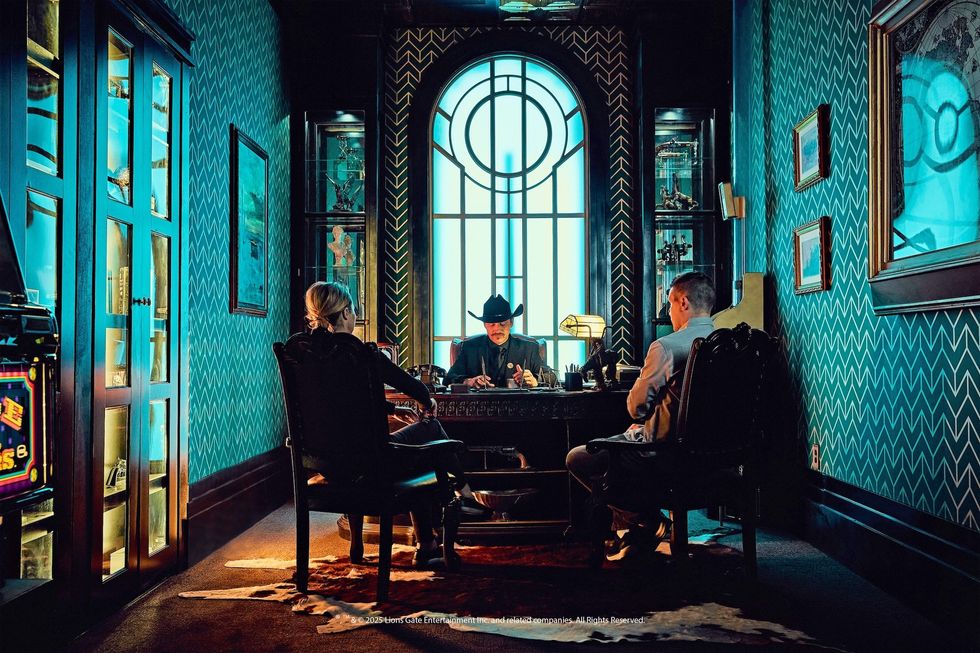 Two people sit facing a cowboy-hatted man in an ornate, blue-patterned office.