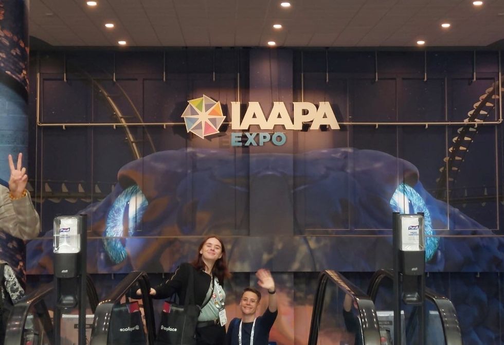 Two people smiling and waving at the IAAPA Expo entrance.
