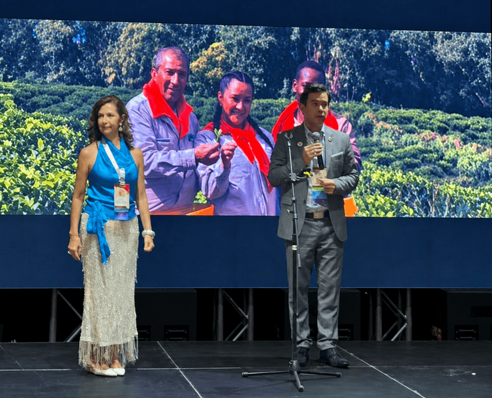 Two people stand on stage, one speaking, with a nature backdrop display.