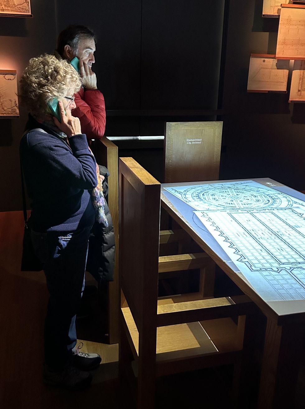 Two people viewing architectural plans on a lit table, holding audio guides.