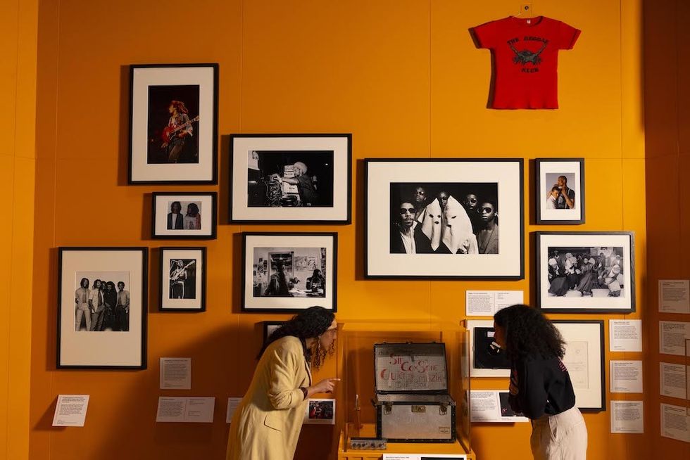 Two people viewing framed photos and memorabilia on an orange gallery wall.