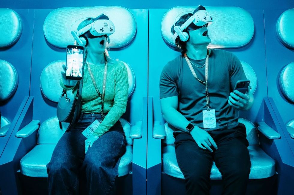 Two people wearing VR headsets, sitting in illuminated chairs, holding smartphones.