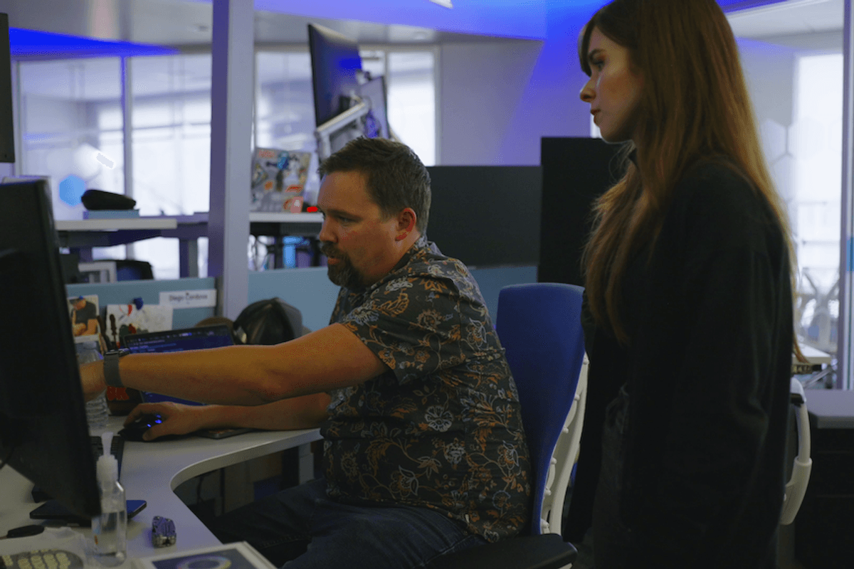 Two people working at a desk, one seated pointing at a computer screen.