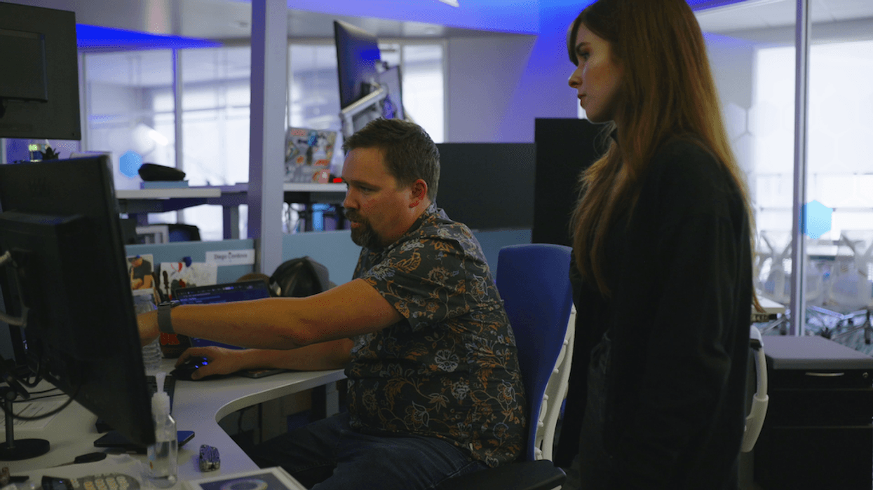 Two people working at a desk, one seated pointing at a computer screen.