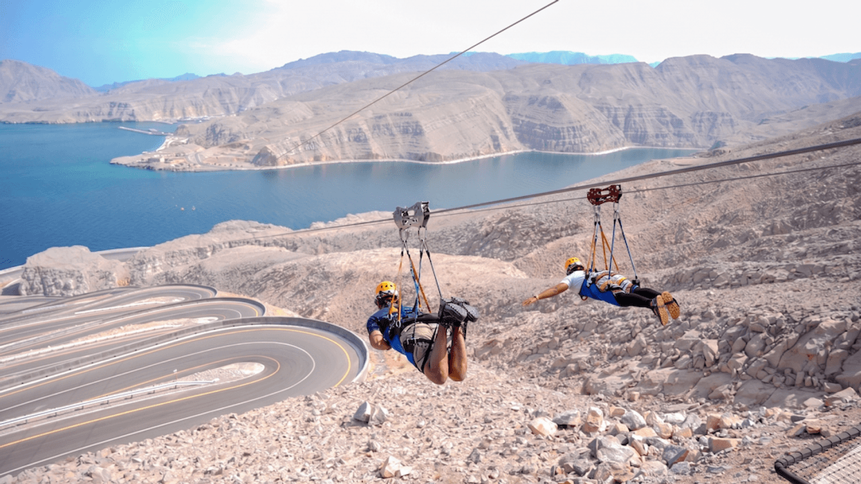 Two people ziplining over rocky terrain with winding road and sea in the background.