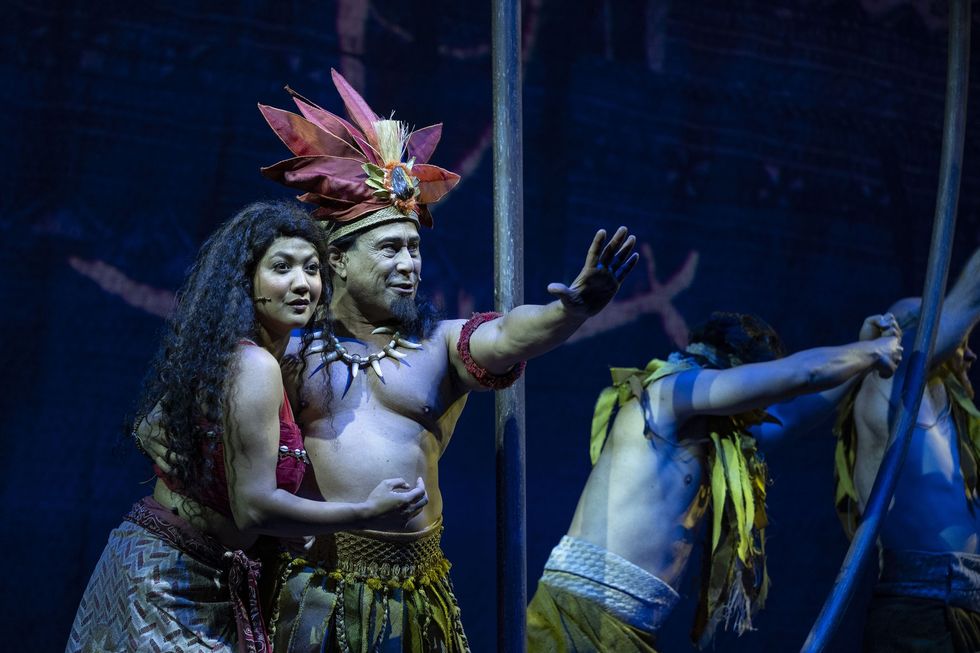 Two performers in tribal costumes on stage, one extending an arm, with a dark backdrop.