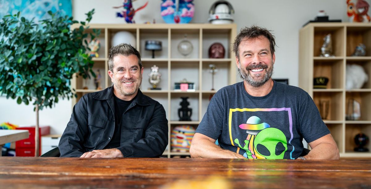 Two smiling men at a wooden table with colorful shelves in the background.