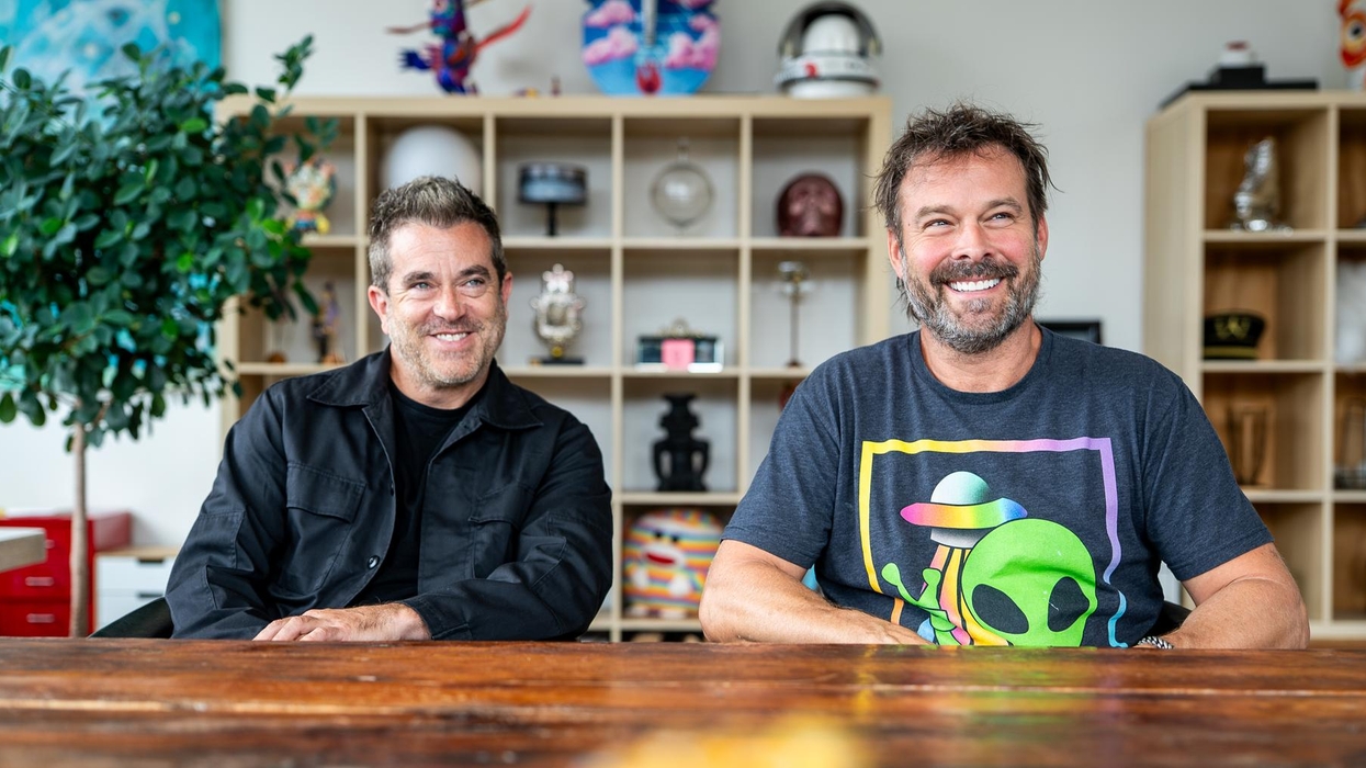 Two smiling men at a wooden table with colorful shelves in the background.