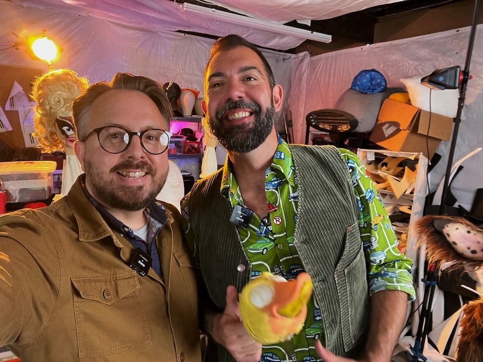 Two smiling men in a colorful, cluttered workshop with costumes and props.