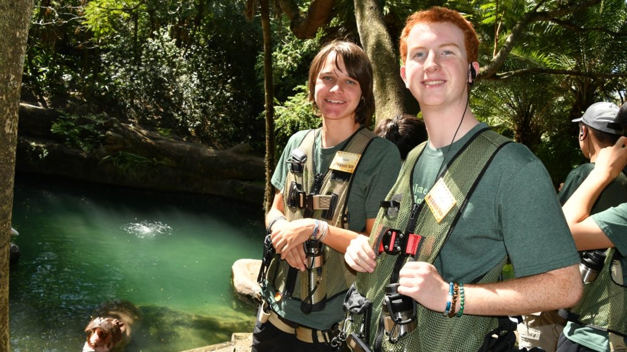 Two smiling visitors in safety harnesses stand beside a green pool in a lush tropical setting.