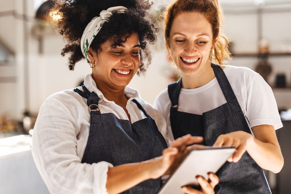 Two smiling women in aprons looking at a tablet in a kitchen.