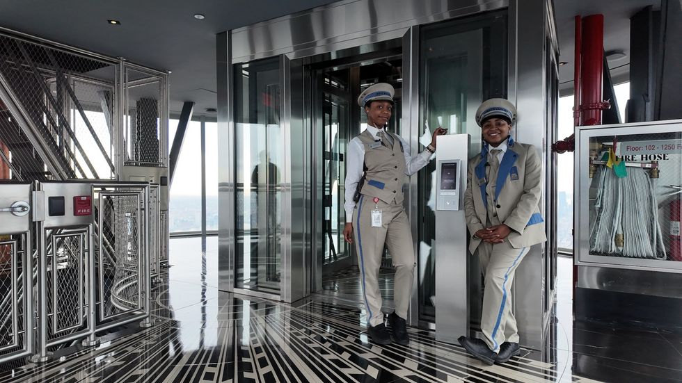 Two uniformed elevator operators smiling beside a glass-walled elevator.